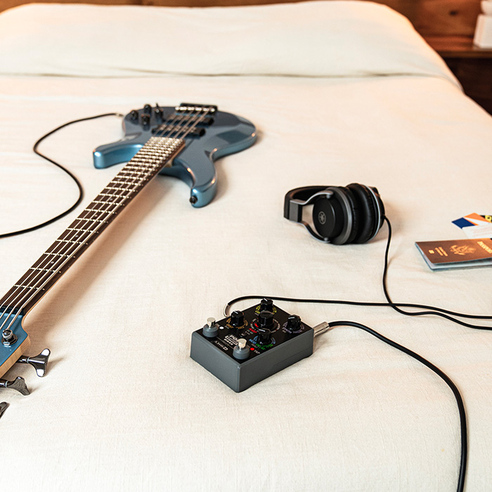 On a hotel bed lies a passport and a POD Express Bass plugged into bass and headphones.