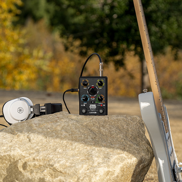 POD Express Black and headphones on a rock outdoors with a guitar leaning against the rock